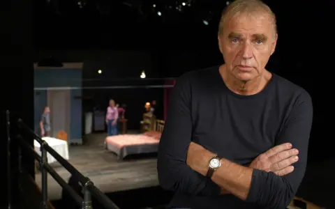 Dawid Głowacki, reżyser teatralny, z rękami skrzyżowanymi na piersi, na tle sceny z łóżkiem i ludźmi.