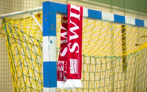 Czerwono-biały szalik z napisem "ŚWIDNICA" wisi na niebiesko-białej bramce do piłki ręcznej z żółtą siatką.