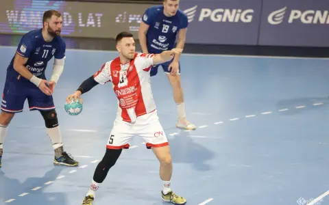 Kamil Adamski w akcji, rzuca piłkę do bramki. Zawodnik w białej koszulce z czerwonymi pasami i numerem 5.
