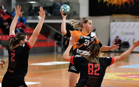 Piłka ręczna: zawodniczka z numerem 21 w pomarańczowej koszulce rzuca do bramki, broniona przez dwie rywalki w czarnych strojach. Łukasz Warchoł uchwycił ten dynamiczny moment.