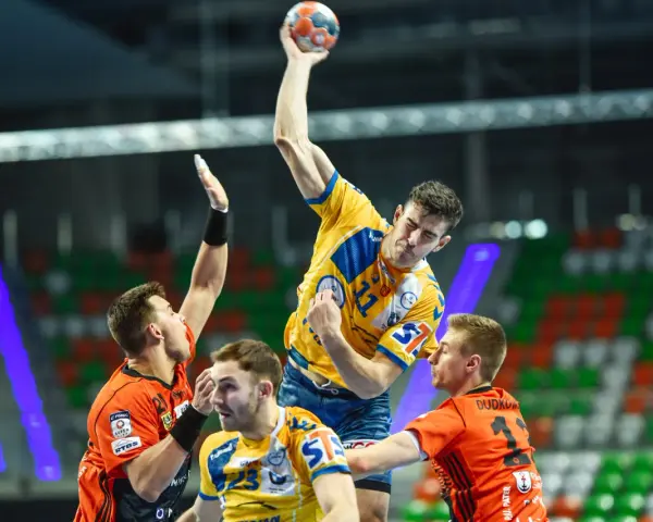Gracz w żółtej koszulce rzuca piłkę do bramki, broniony przez zawodników w pomarańczowych strojach. Emocjonujący moment z meczu final four handball.