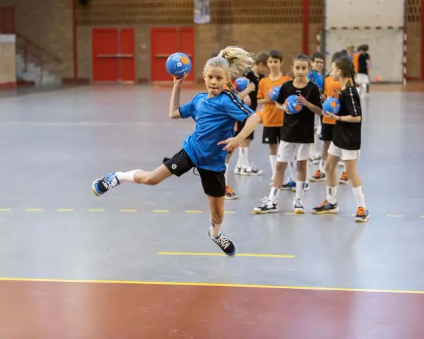 Dziewczynka w skoku wykonuje kozłowanie w piłce ręcznej, podczas gdy inni gracze czekają na swoją kolej.