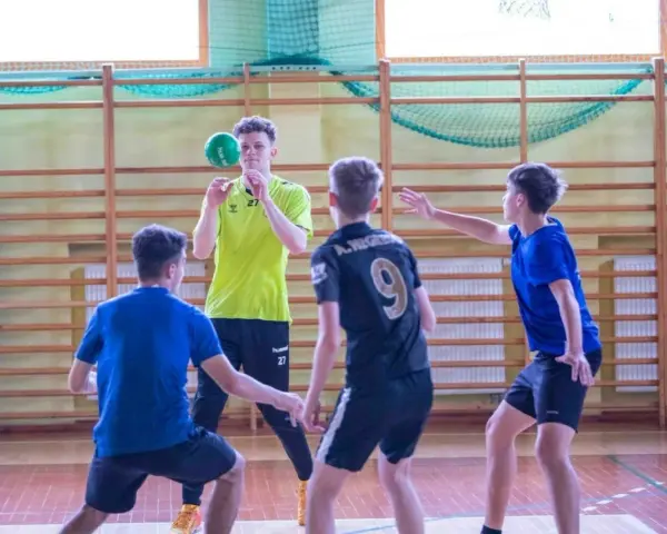 Cezary Surgiel w żółtej koszulce rzuca piłkę do ręcznej, otoczony przez kolegów z drużyny na sali gimnastycznej.
