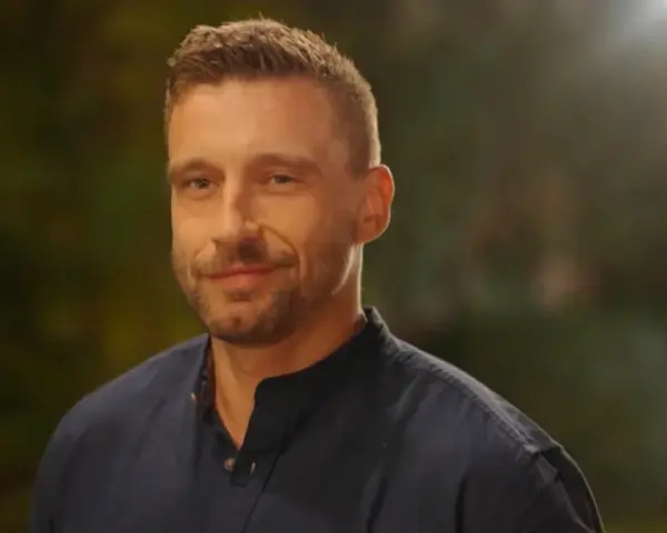 Uśmiechnięty Jakub Wojnowski w granatowej koszuli, w tle rozmyte światła i zieleń.