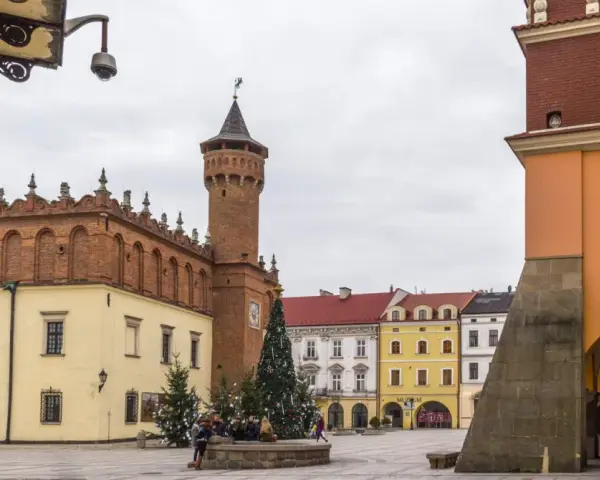 Rynek w Tarnowie zimą, z choinką i ludźmi przy fontannie. W tle zabytkowe kamienice i wieża ratuszowa.