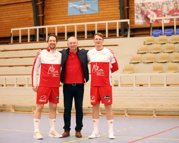 Damian Kędziora z dwoma zawodnikami w strojach sportowych, na tle trybun hali sportowej.