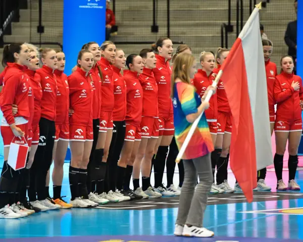 Drużyna piłki ręcznej kobiet Polski w czerwonych strojach stoi w rzędzie, trzymając polską flagę.