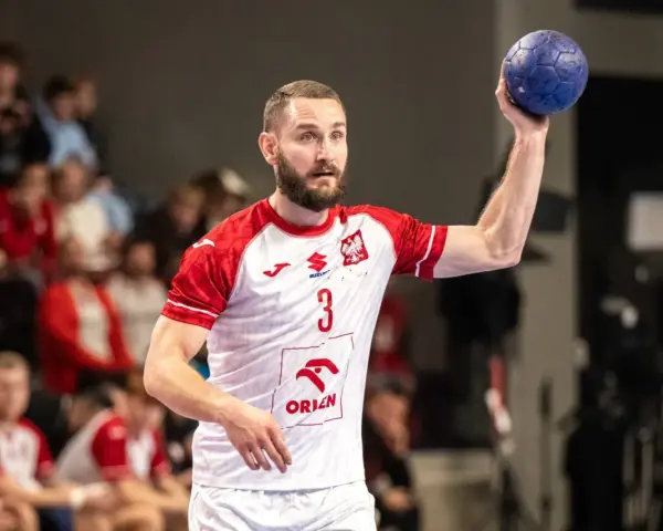 Gracz w białej koszulce z czerwonymi rękawami, numerem 3 i godłem Polski, trzyma niebieską piłkę ręczną, gotowy do rzutu.