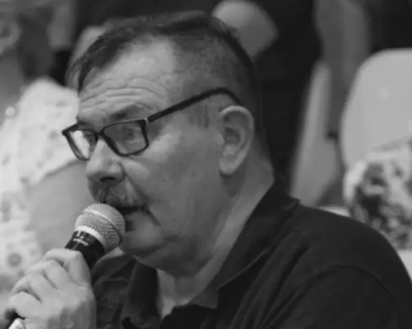 Krzysztof Janicki w okularach i z wąsami mówi do mikrofonu, w tle widać ludzi.