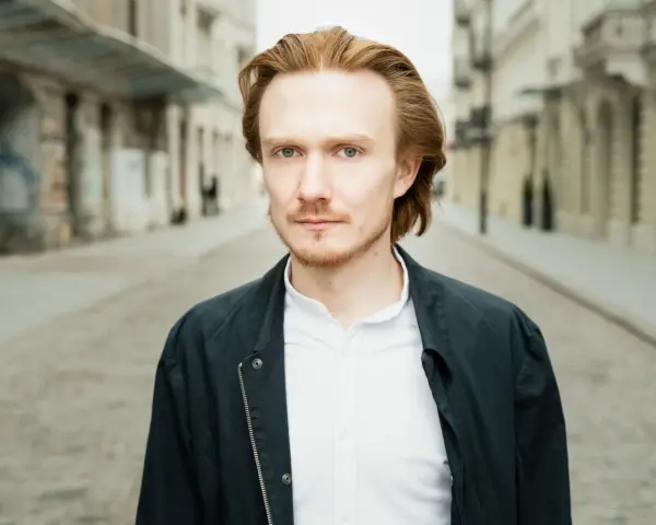 Bartosz Bednarski na tle miejskiej ulicy, z rudymi włosami i zielonymi oczami, w białej koszuli i ciemnej kurtce.