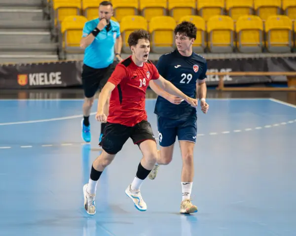 Jakub Smoliński w czerwonej koszulce z numerem 14 biegnie z piłką ręczną, obok zawodnik w granatowej koszulce z numerem 29.