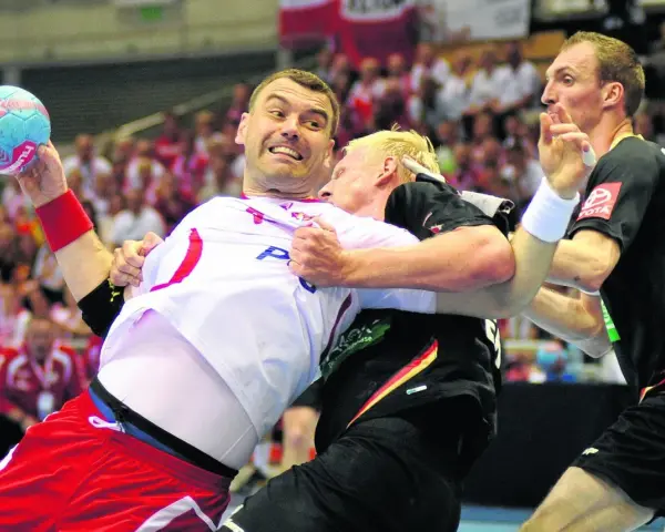Gracz w białej koszulce z piłką ręczną próbuje wykonać rzut karny, podczas gdy przeciwnik próbuje go zatrzymać.