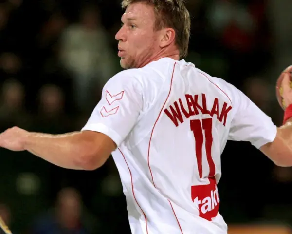 Damian Wleklak, numer 11, w białej koszulce z czerwonymi detalami, przygotowuje się do rzutu piłką ręczną.