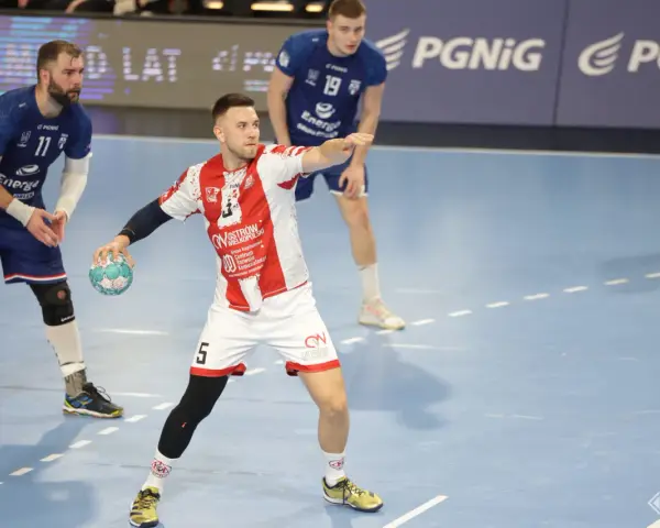 Kamil Adamski w akcji, rzuca piłkę do bramki. Zawodnik w białej koszulce z czerwonymi pasami i numerem 5.