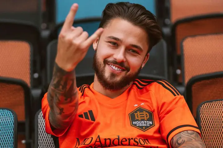 Uśmiechnięty Adam Kowalczyk w koszulce Houston Dynamo FC pozdrawia gestem rock and rolla, siedząc na trybunach.