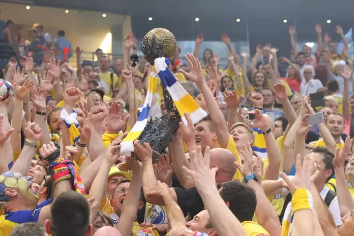 Tłum świętuje zwycięstwo, unosząc puchar. Kibice w żółto-niebieskich barwach, jak na meczach kielce liga mistrzów, cieszą się z sukcesu.
