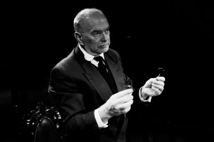 Piotr Wyszomirski w eleganckim garniturze, trzymający w dłoniach kieliszek i coś na patyczku.