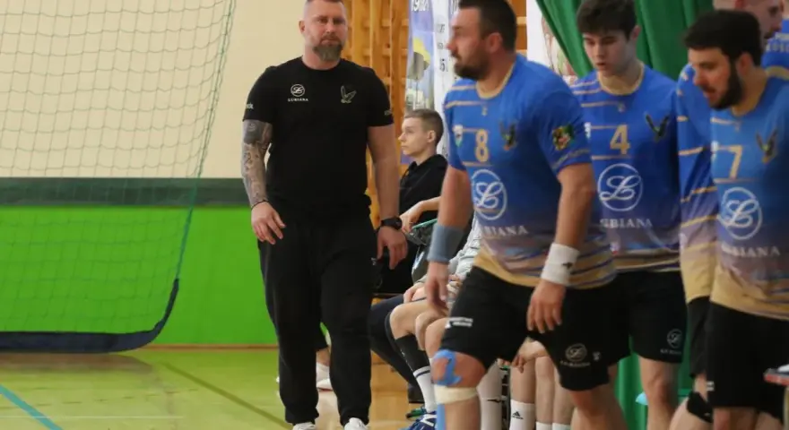 Maciej Reichel, trener, obserwuje mecz piłki ręcznej. Zawodnicy w niebieskich koszulkach czekają na swoją kolej.