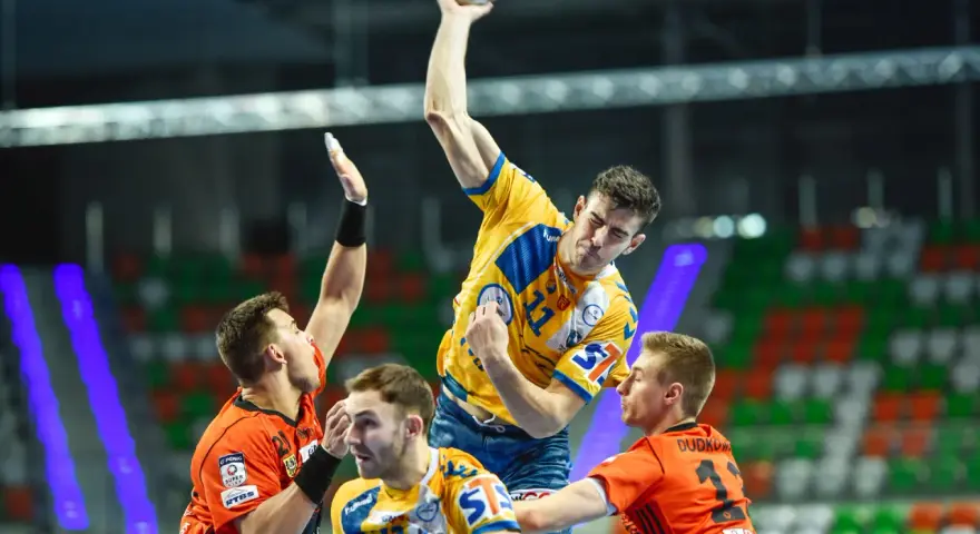 Gracz w żółtej koszulce rzuca piłkę do bramki, broniony przez zawodników w pomarańczowych strojach. Emocjonujący moment z meczu final four handball.