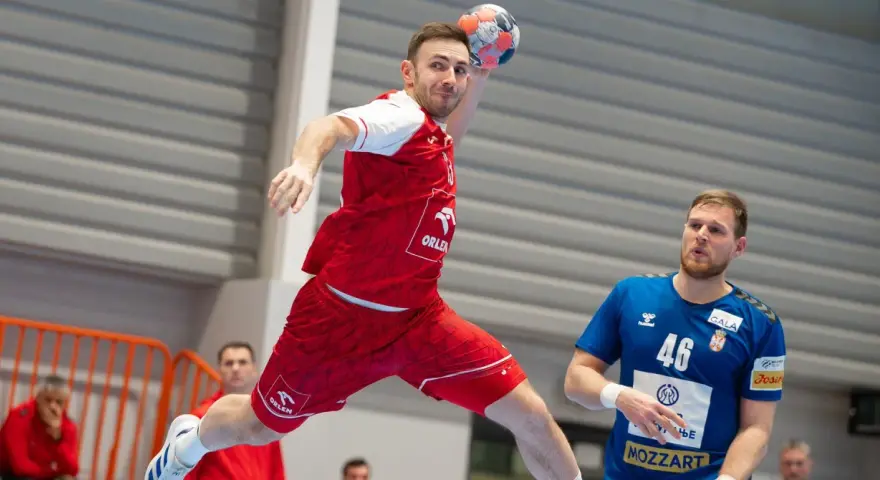 Piłkarz ręczny w skoku do rzutu, w czerwonej koszulce z logo ORLEN. Obok niego stoi zawodnik w niebieskiej koszulce z numerem 46. To reprezentacja Serbii w piłce ręcznej mężczyzn.