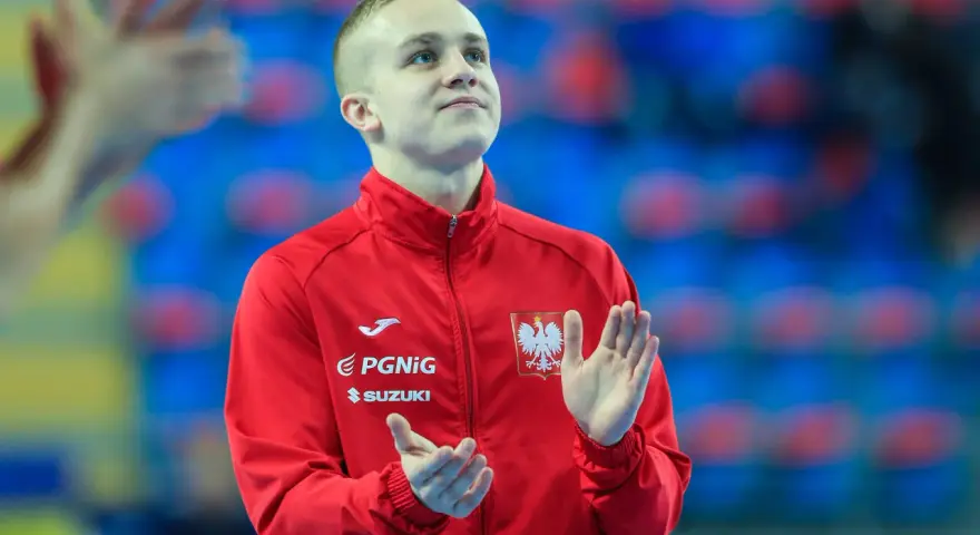 Adam Jędraszek w czerwonej kurtce z orłem, z dłońmi uniesionymi w geście radości lub powitania.
