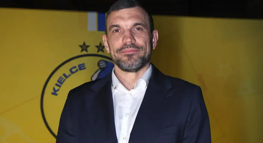 Michał Jurecki w garniturze na tle żółtej ściany z logo "Kielce".