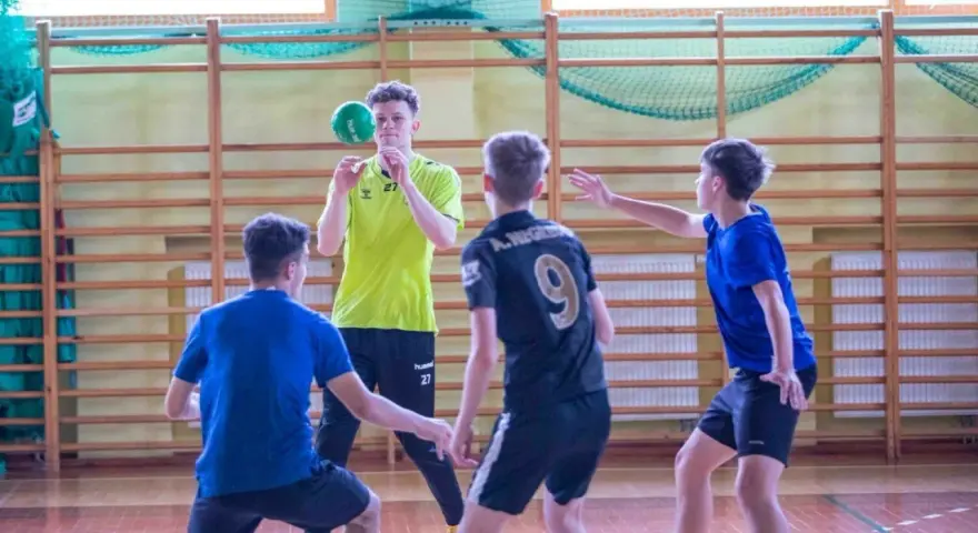 Cezary Surgiel w żółtej koszulce rzuca piłkę do ręcznej, otoczony przez kolegów z drużyny na sali gimnastycznej.