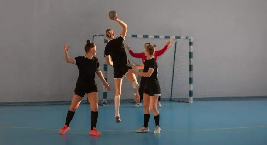 Piłkarki ręczne w akcji. Jedna z nich skacze do rzutu, podczas gdy koleżanki z drużyny i przeciwniczka obserwują. To dynamiczna gra pasywna w piłce ręcznej.