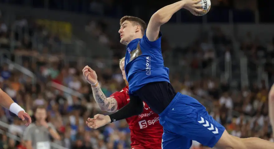 Magnus Landin Jacobsen w skoku do rzutu piłką ręczną, w niebieskim stroju, z widownią w tle.