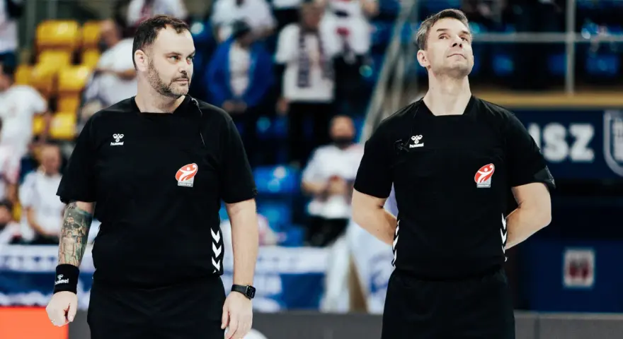 Dwóch sędziów w czarnych koszulkach z logo Hummel i emblematem związku sportowego, gotowych do rozpoczęcia meczu ligi piłki ręcznej.