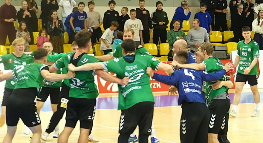 Krystian Woźniak i jego drużyna świętują zwycięstwo. Zawodnicy w zielonych strojach przytulają się na boisku, a na trybunach widać kibiców.