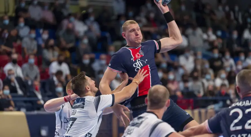 Dainis Kristopans w skoku rzuca piłką do bramki, otoczony przez obrońców.