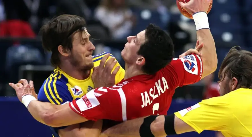 Zacięta walka w meczu polska szwecja piłka ręczna. Zawodnicy walczą o piłkę, jeden w czerwonej koszulce z napisem Jaszka, drugi w żółtej.