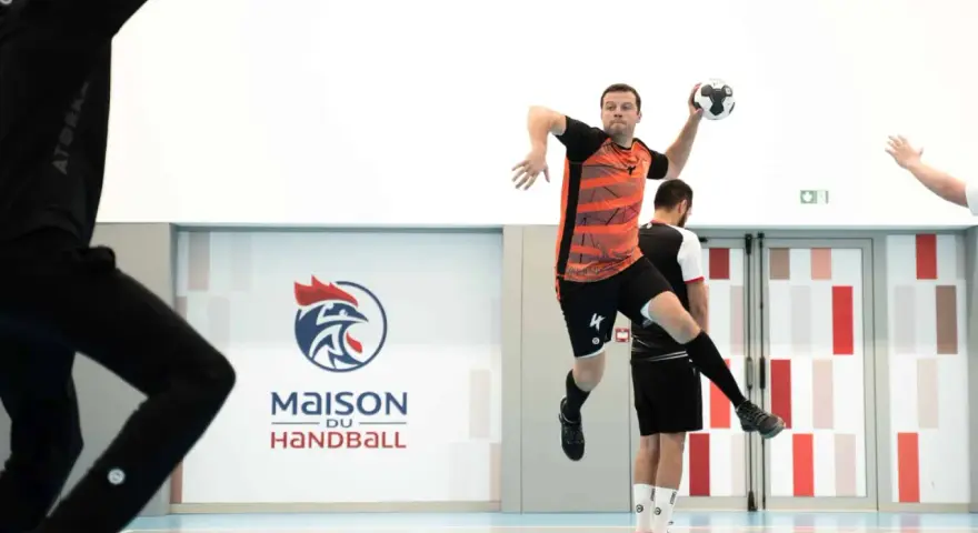 Piłkarz ręczny, niczym szczypiornista, wyskakuje do rzutu. W tle logo "Maison du Handball".