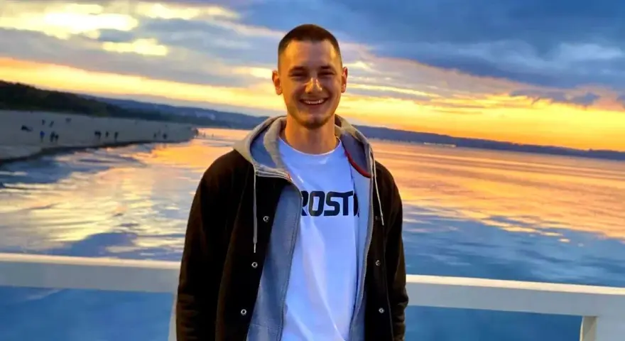 Uśmiechnięty Krzysztof Komarzewski na molo o zachodzie słońca. W tle plaża i morze.