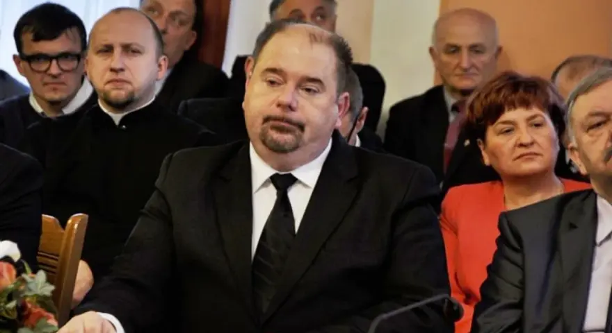 Jarosław Paluch w czarnym garniturze siedzi przy stole, otoczony innymi osobami.