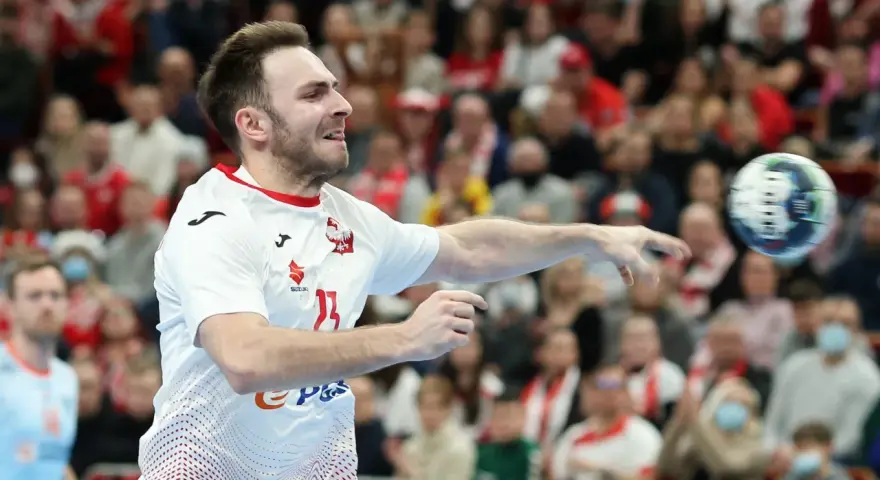 Piłkarz ręczny Polski w akcji, rzuca piłkę. W tle widać trybuny z kibicami, którzy dopingują swoją drużynę w meczu przeciwko Iranowi.