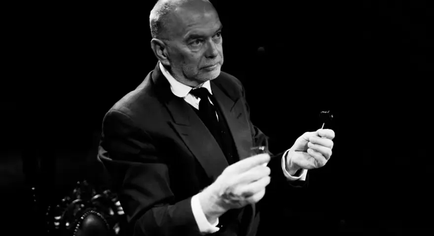 Piotr Wyszomirski w eleganckim garniturze, trzymający w dłoniach kieliszek i coś na patyczku.
