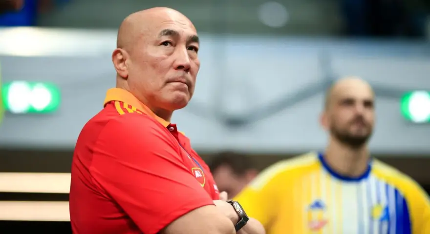 Skupiony trener Vive Kielce w czerwonej koszulce z pomarańczowym kołnierzem, z rękami skrzyżowanymi na piersi.