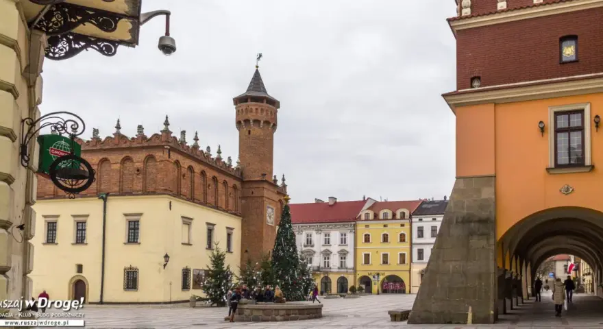 Rynek w Tarnowie zimą, z choinką i ludźmi przy fontannie. W tle zabytkowe kamienice i wieża ratuszowa.