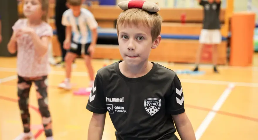 Chłopiec z zabawką na głowie, w koszulce KS Bysstra, podczas zajęć sportowych.
