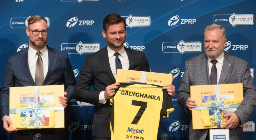 Trzech mężczyzn w garniturach prezentuje koszulkę z napisem "Galychanka" i numerem 7. Obok nich żółte pudełka z napisami "Poland Ukraine".
