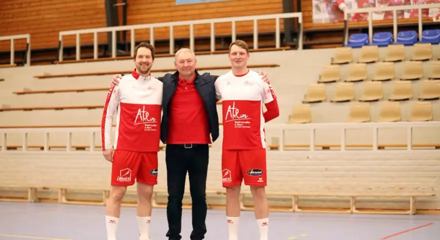 Damian Kędziora z dwoma zawodnikami w strojach sportowych, na tle trybun hali sportowej.