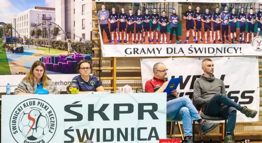 Na banerze widnieje drużyna SKPR Świdnica. Dwie kobiety siedzą za stołem z piłką ręczną.