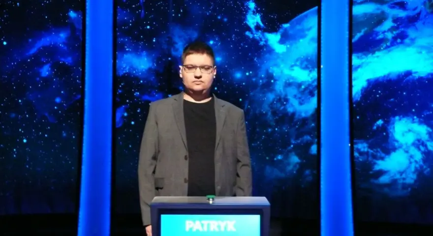 Patryk w szarym garniturze stoi przed niebieskim tłem z gwiazdami.