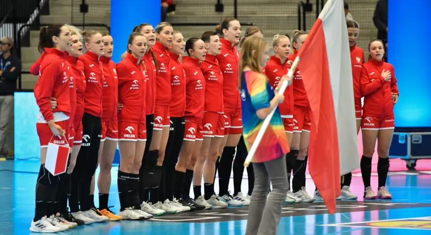 Drużyna piłki ręcznej kobiet Polski w czerwonych strojach stoi w rzędzie, trzymając polską flagę.