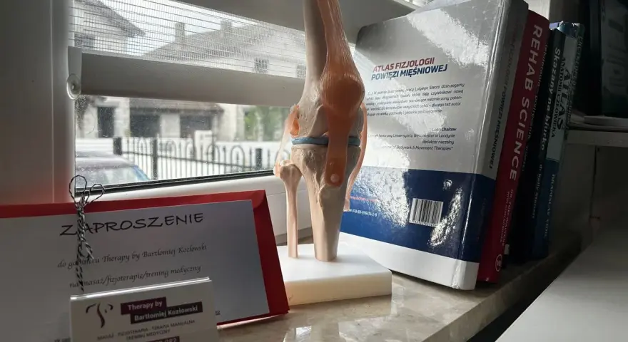 Model stawu kolanowego obok zaproszenia na terapię od Bartłomieja Kozłowskiego i książek o fizjoterapii.