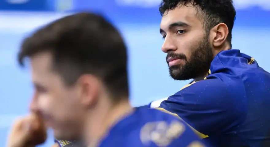 Hassan Kaddah, z brodą i kręconymi włosami, w niebieskiej bluzie z żółtymi detalami, spogląda w bok.