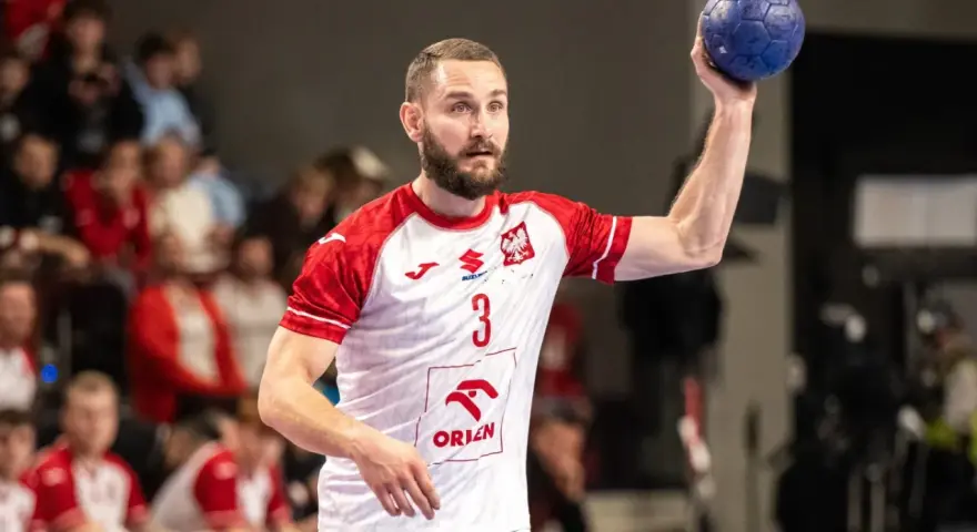 Gracz w białej koszulce z czerwonymi rękawami, numerem 3 i godłem Polski, trzyma niebieską piłkę ręczną, gotowy do rzutu.