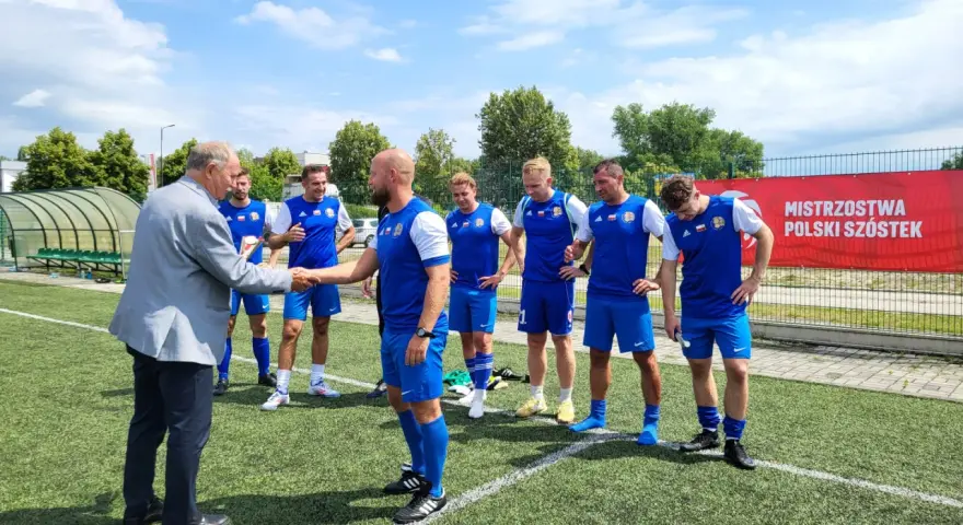 Starszy pan wita drużynę Grunwald Ruda Śląska na boisku. Piłkarze w niebieskich strojach czekają na rozpoczęcie meczu.